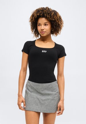 Νεαρή γυναίκα φοράει μαύρο t-shirt GAP με τετράγωνη λαιμόκοψη και μίνι φούστα σε ασπρόμαυρο καρό, στέκεται μπροστά σε μονόχρωμο φόντο.