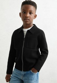 Veste noire à fermeture éclair avec deux poches avant et un col, associée à un jean en denim bleu et un t-shirt blanc en dessous. Texture lisse.