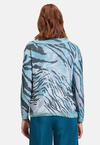 Stickad tröja med ett mönster av blå och grå tigerstrimmor, med krage och strukturerad yta, i kombination med turkosa vida byxor.