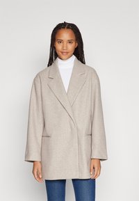 Hellbeige übergroße Blazer aus strukturiertem Stoff mit Reverskragen und zwei vorderen Taschen, getragen über einem weißen Rollkragenpullover.