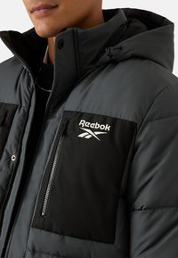 Reebok pufferjack in donkergrijs met zwarte accenten. Heeft een borstzak met rits en logodetail. Gemaakt van een getextureerd, doorgestikt materiaal.