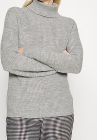 Pull à col roulé gris clair avec un motif en maille texturée et des poignets côtelés. Associé à un pantalon à motif pied-de-poule.