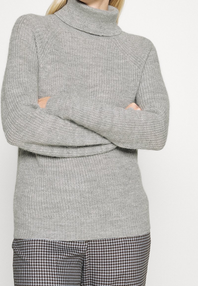 Pull à col roulé gris clair avec un motif en maille texturée et des poignets côtelés. Associé à un pantalon à motif pied-de-poule.