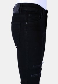 Zwarte denim jeans met een slim fit, verwerkte kniestukken en een leren label op de tailleband. Zijaanzicht toont het ontwerp van de zakken.