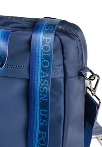 Mörkblå väska i glänsande syntetmaterial, med dragkedjestängning, tjockt strukturerat band med gradient i blå toner och metallbeslag.