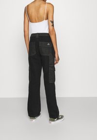 En person i svarta cargobyxor, en vit tanktop med tunna axelremmar, och gröna Vans-sneakers, stående mot en ljus enfärgad bakgrund.