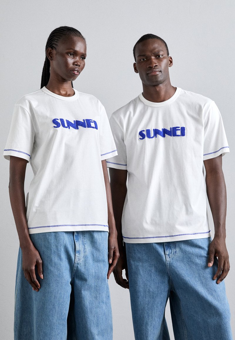 Λευκά βαμβακερά μπλουζάκια με μπλε κεντητό λογότυπο "SUNNEI", με αντίθετη μπλε ραφή στα μανίκια και την κάτω πλευρά, συνδυασμένα με ανοιχτόχρωμα τζιν παντελόνια.