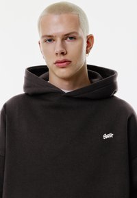 Dunkelbrauner Kapuzenpulli mit Kordelzug, ausgestattet mit einem weißen Logo auf der linken Brust. Glatte Textur mit einem lässigen Schnitt.