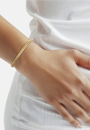 Poignet portant un bracelet fin en spirale dorée reposant sur un haut en tissu blanc texturé.