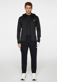 Svart huvorjacka med dragkedja, bärs över en svart polo. Kombinerad med svarta byxor och vita sneakers. Gummisula och slimmad design.