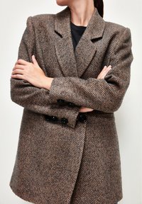 Nainen, jolla on kädet ristissä, pukeutuneena ruskeaan ja mustaan herringbone-kuosiset takki mustan paidan päällä, päänsä käännettynä sivulle.