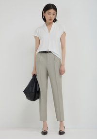 Donna che indossa una blusa bianca a maniche corte, pantaloni beige a vita alta, cintura nera, sabot neri con tacco a blocco, mentre tiene una grande borsa nera.