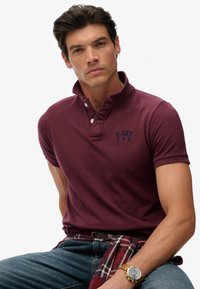 Bordeauxrode poloshirt met een button-down kraag en een klein blauw logo op de borst. Gecombineerd met een geruit overhemd, vastgeknoopt rond de taille.