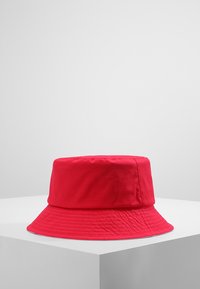 Cappello a secchiello rosso realizzato in morbido tessuto, caratterizzato da una larga visiera, una texture liscia e una forma cilindrica e strutturata con cuciture visibili.