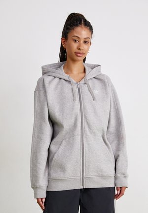 Giovane donna con capelli intrecciati, che indossa un felpa grigia chiara con zip e pantaloni neri, in piedi di fronte a uno sfondo bianco semplice.