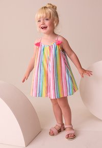 Kleurrijke gestreepte jurk met roze kwastaccenten, mouwloos ontwerp en een uitlopende silhouet. Gecombineerd met lichtroze sandalen.