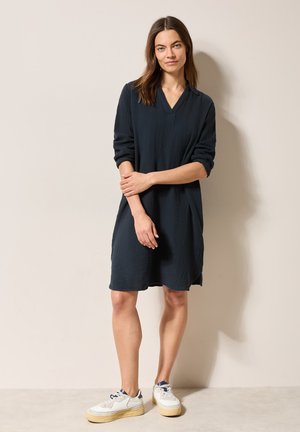 Mörkblå oversized klänning med v-ringning och långa ärmar, gjord av ett texturerat tyg, tillsammans med vita sneakers. En enkel beige bakgrund.