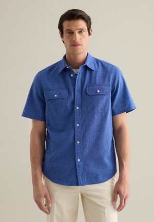 Camisa de manga corta, con botones, en tejido texturizado de color azul, con dos bolsillos en el pecho y botones blancos, combinada con pantalones beige claros.