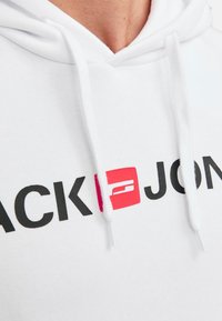 Hvid hoodie med kængurulomme, justerbare snore og et stort sort logo med rød accent på brystet. Blødt stof.