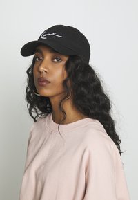 Svart keps med vit kursiv logotyp, använd med en ljusrosa sweatshirt. Har strukturerad tyg och böjd skärm. Avslappnad passform.