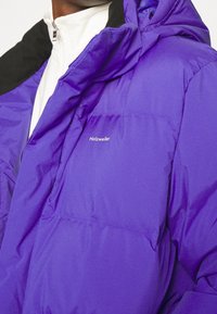 Osoba v purpurové péřové bundě s logem "Holzweiler" přes bílou zipovou mikinu, ukazující horní část hrudníku a ramena.