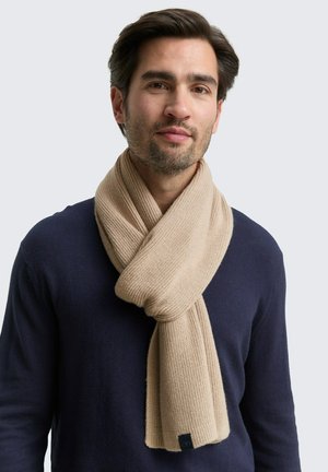 Beige knitted scarf wrapped around the neck, with a ribbed texture. Tags visible at the end. Worn over a navy sweater.