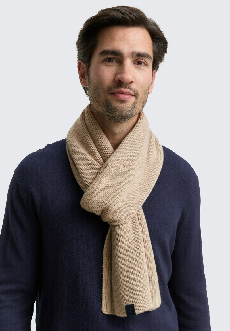 Beige gebreide sjaal om de nek gewikkeld, met een geribd patroon. Labels zichtbaar aan het uiteinde. Draagt over een marineblauw trui.
