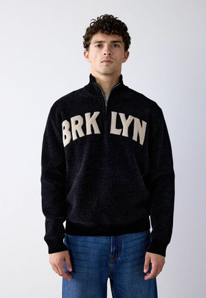 Young man with curly hair wearing a black zip-up sweater with "BRKLYN" in beige letters and blue jeans, standing against a plain white background.
