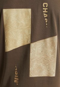 Tissu marron avec deux motifs carrés beiges comportant des lignes topographiques abstraites blanches et un texte beige partiellement visible aligné verticalement.