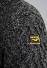 Suéter gris de punto con un patrón de trenzas, con un parche de marca amarillo etiquetado como "PME Legend American Classic" en el hombro izquierdo.