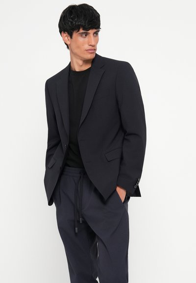 Blazer navy de tela suave, con un corte entallado, solapas de muesca y dos bolsillos frontales, llevado sobre una camiseta negra y pantalones oscuros.