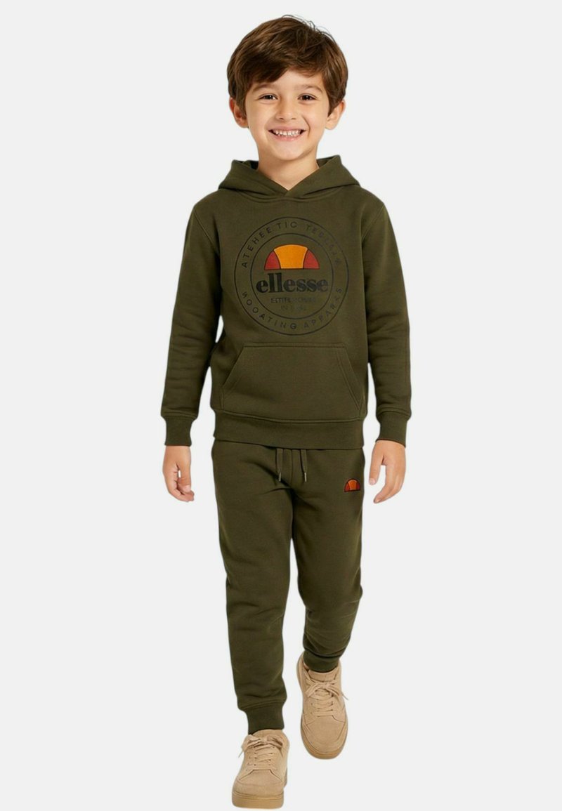 Garçon souriant marchant, portant un sweat à capuche vert olive et un pantalon de jogging assorti avec un logo, des baskets beiges, sur un fond blanc uni.