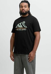Sort t-shirt med korte ærmer, der har et bjergmotiv i grøn og guld tekst, lavet af bomuld med en afslappet pasform. Bæres med lysegrå bukser.
