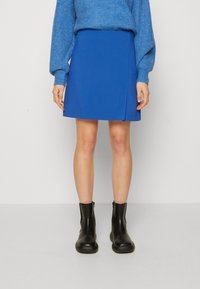 Niebieska mini spódniczka o prostym kroju, z bocznym rozcięciem i gładką fakturą, w połączeniu z czarnymi botkami sięgającymi kostki. Widoczny jest także przytulny niebieski sweter.