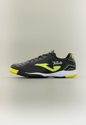 Chaussure de football en salle noire et jaune avec semelle blanche, design à lacets et texte "Toledo" sur le côté sur un fond uni.