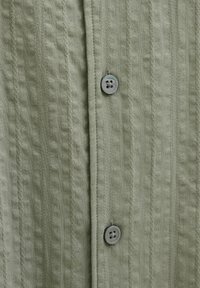 Košulja s gumbima napravljena od teksturirane, svijetlozelene tkanine s vertikalnim prugama i okruglim detaljem s četiri rupe za gumbe.