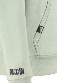 Lichtgroene sweatshirt met geribbelde manchetten en onderrand, voorzien van marineblauwe borduursels van "MOIN" en een ankerdetail. Zakken aan de zijkant met een logo-label.