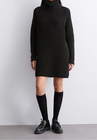 Robe en tricot noir avec un col roulé haut et des manches longues. Présente un motif texturé, coupe mi-genou, portée avec des chaussettes noires montantes et des chaussures.