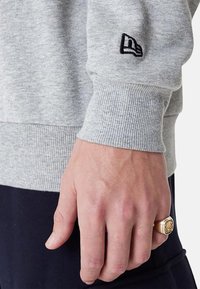 Sudadera gris con puños y dobladillo acanalados, con un logo bordado en negro en la manga. La mano lleva un anillo dorado con un patrón decorativo.