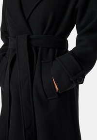 Accappatoio in misto wool nero con design avvolgente, dotato di cintura da annodare, tasche laterali e polsini con bottoni. Texture morbida, vestibilità strutturata.