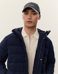 Ung mann iført navy puffer-jakke over en kremfarget ribbestrikket genser med glidelås og mørk grå baseballcap, stående mot en enkel lys bakgrunn.