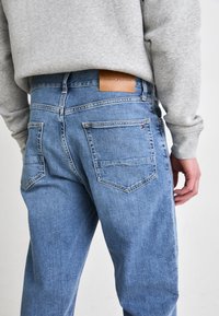Light blue denim jeans with a straight cut, featuring a leather back patch, two rear pockets, and subtle texture details.