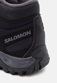 Melnas pārgājienu zābaki ar mīkstu apkaklīti, teksturētu audumu, gumijas zoli un pelēkiem akcentiem. Uz sāniem ir "Salomon" logo.