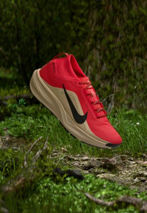 Sarkana un bēša Nike skriešanas apavi ar melnu swoosh peldo virs meža zemes ar augiem un akmeņiem.