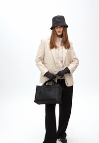 Modelka má na sobě béžový blazer, černé rukavice a černý klobouk typu bucket, přičemž drží malou černou tašku na pozadí z čistě bílé.