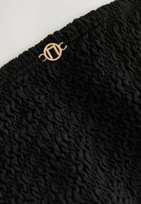 Melnas teksturētas blūzes ar smocka dizainu un zelta apļa detaļu jostasvietā, izceļot tā sarežģītās rakstu formas.