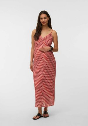 Femme aux longs cheveux foncés portant une robe maxi sans manches rayée rouge et des sandales noires, debout avec une main sur son ventre.