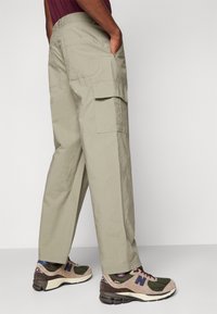 Světlé khaki cargo kalhoty s bočními kapsami, volný střih a jemná textura, kombinované s béžovými a zelenými teniskami s logem.