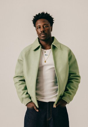 Mann steht mit den Händen in den Taschen, trägt eine hellgrüne Jacke, ein weißes Hemd, goldene Halsketten und dunkle Jeans vor einem einfarbigen Hintergrund.