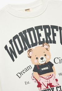 Sudadera de color crema claro con un oso de dibujos animados que lleva una camiseta negra con el texto "WONDER", combinada con pantalones rosas; diseño gráfico audaz.